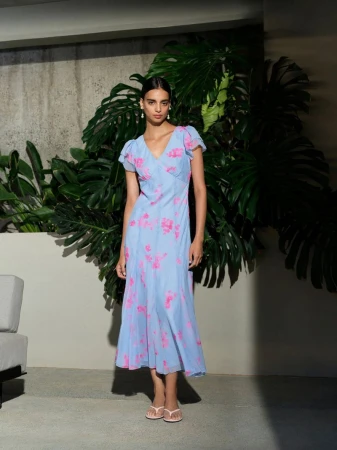 A fair-skinned young woman stands in front of large tropical leaves, modeling a light blue floral dress and pale pink flip-flops.