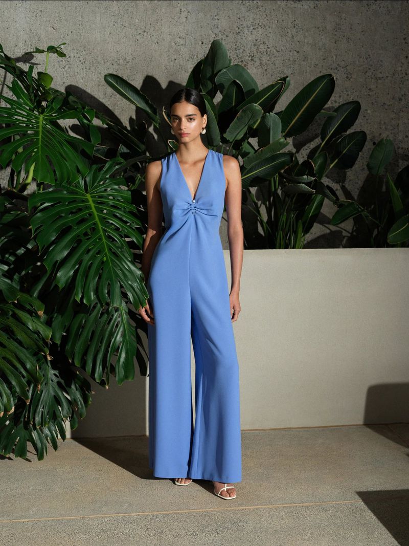 A woman in a blue jumpsuit stands in front of tropical plants against a neutral wall, presenting fashion and style in an indoor setting.