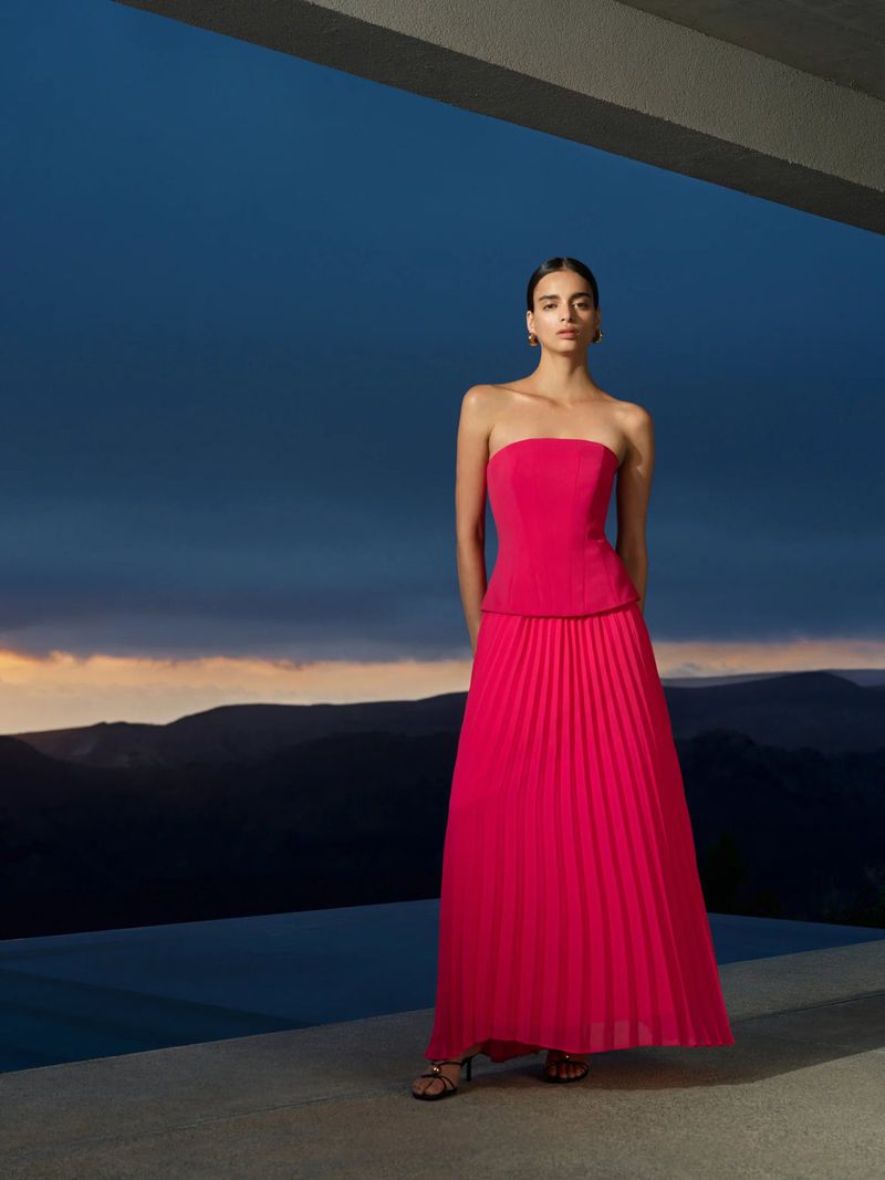 Elegant woman in a strapless pink dress stands on a terrace overlooking mountains under a dark blue sky at dusk, fashion editorial.