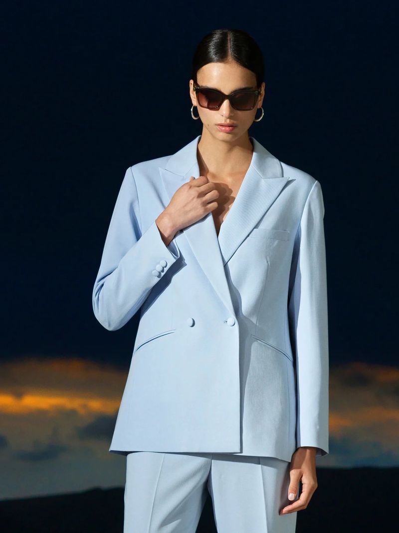 A stylish adult female wearing a baby blue pantsuit and sunglasses poses against a blurred background of dark blue and orange.