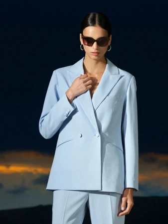A stylish adult female wearing a baby blue pantsuit and sunglasses poses against a blurred background of dark blue and orange.