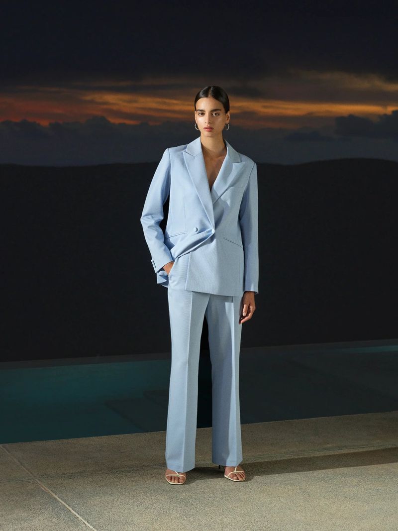 A woman in a light blue pantsuit poses outdoors against a dark wall with orange clouds in the distance, showcasing a fashion-forward aesthetic.