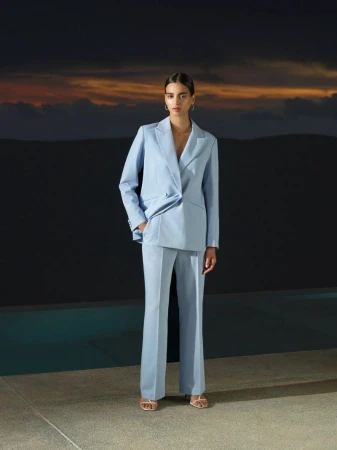 A woman in a light blue pantsuit poses outdoors against a dark wall with orange clouds in the distance, showcasing a fashion-forward aesthetic.