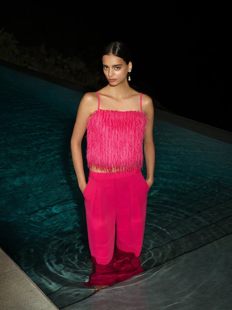 Woman in pink fringe top and pants standing in pool at night. Fashion shot with dark, moody lighting, and serene poolside environment.