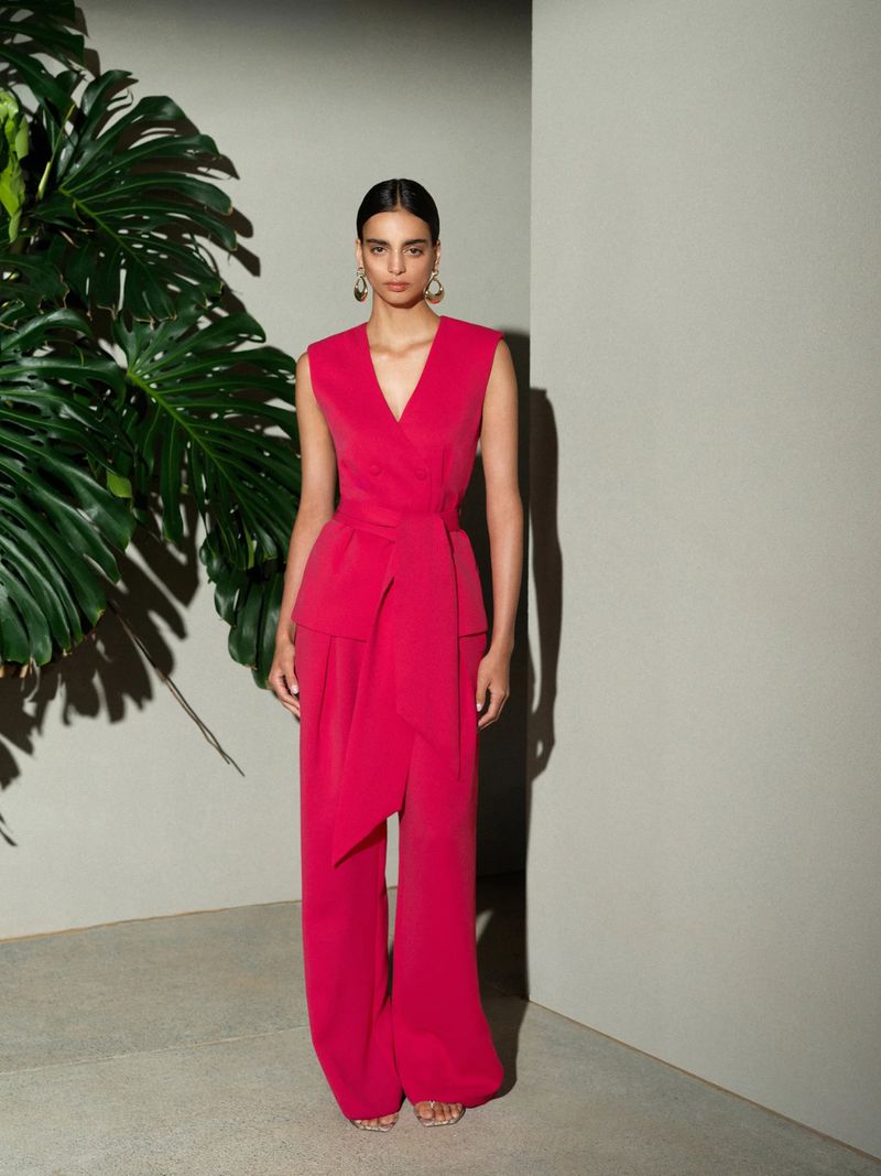 A woman models a vibrant pink wraparound sleeveless top and wide-legged pants in a minimalist studio setting with a large green plant.