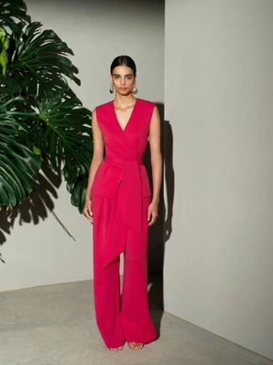 Model in magenta jumpsuit standing beside large green tropical leaves against a neutral grey wall