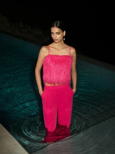 Model standing waist-deep in a swimming pool wearing a bright pink fringed top and matching pink trousers at night.