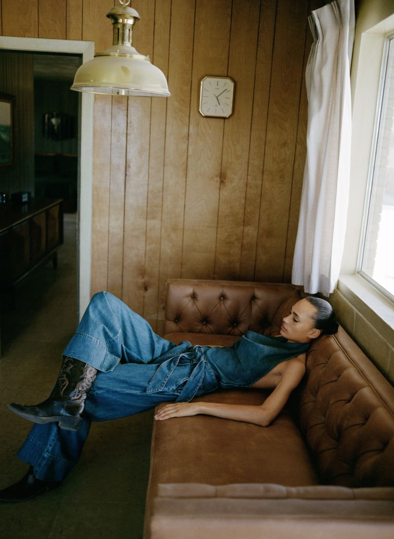 A woman in a denim outfit and cowboy boots reclines on a brown couch in a wood-paneled room with natural light from a nearby window.