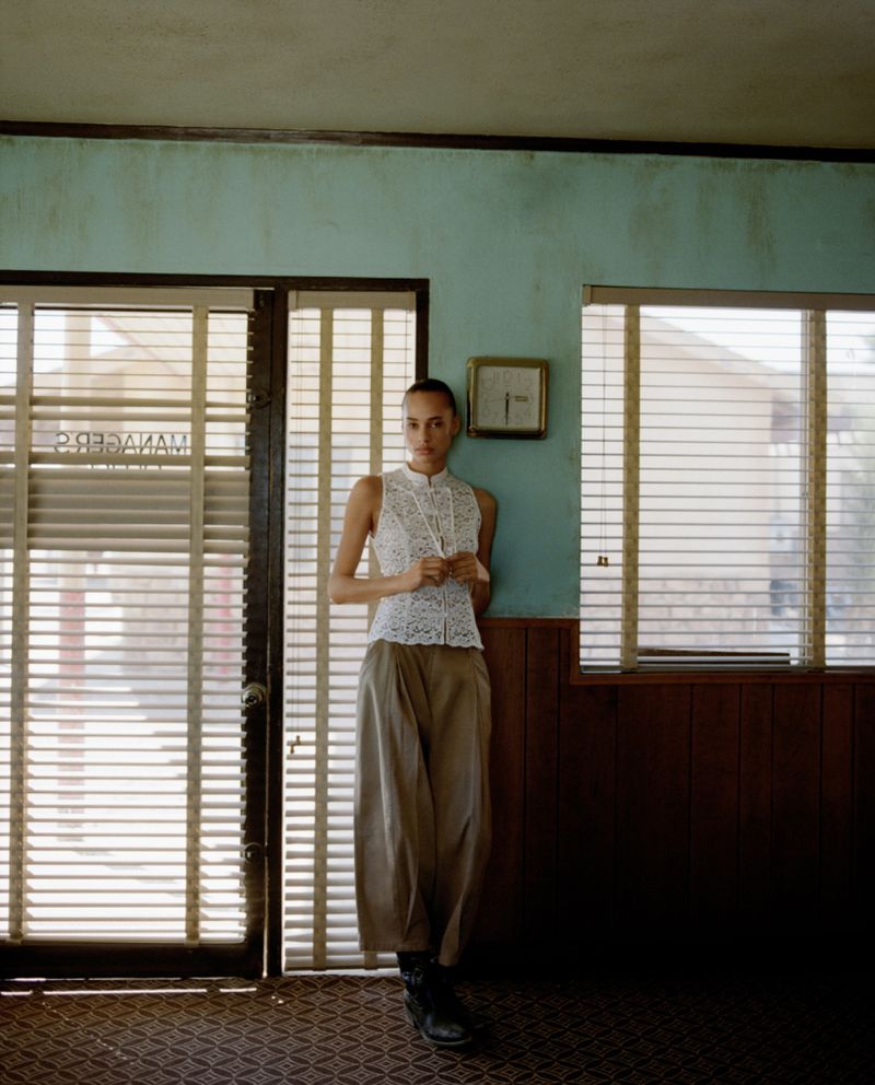 Fashion photo of a slender woman with tied back hair wearing a sleeveless white top and taupe pants, posing in a vintage room with blinds.