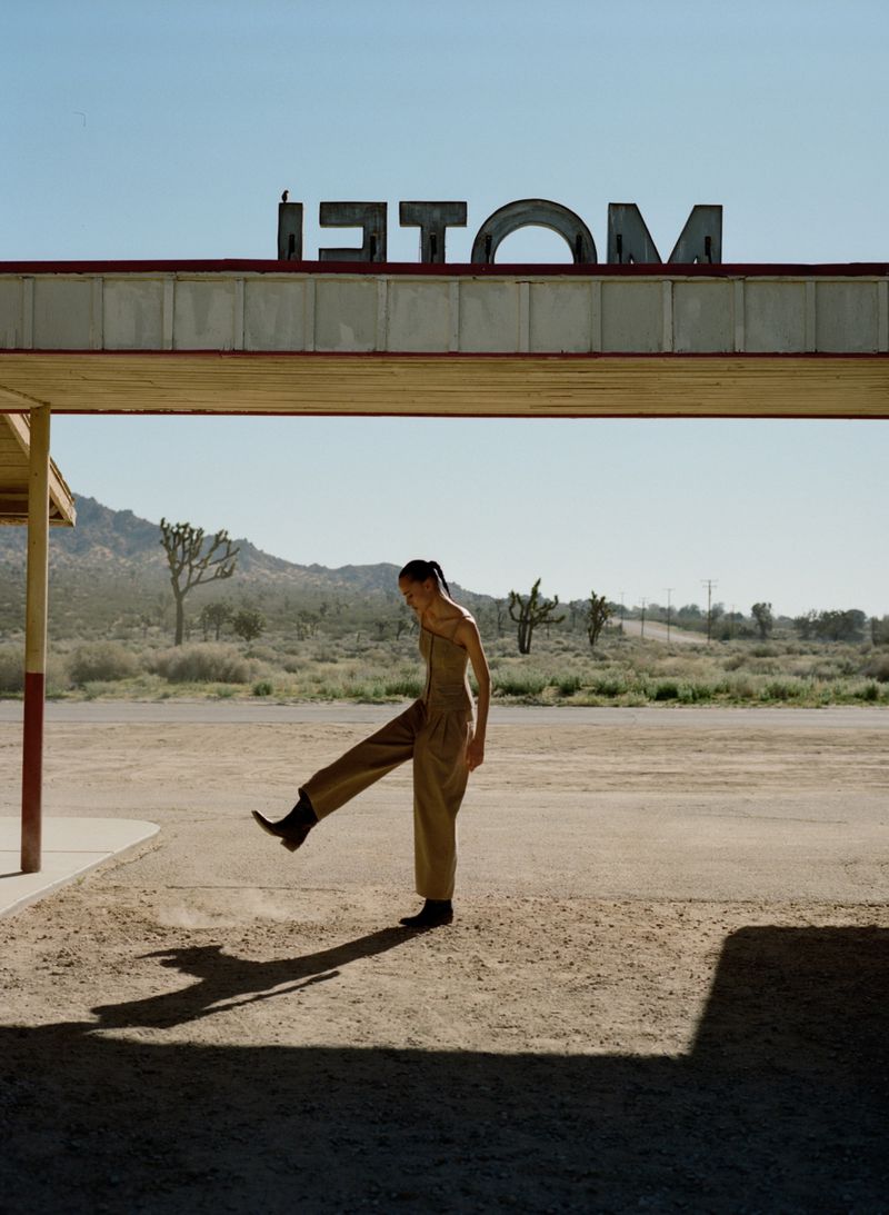 An adult female poses in the desert beneath an old motel sign. She wears a tan jumpsuit and black cowboy boots.