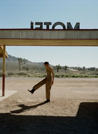 An adult female poses in the desert beneath an old motel sign. She wears a tan jumpsuit and black cowboy boots.