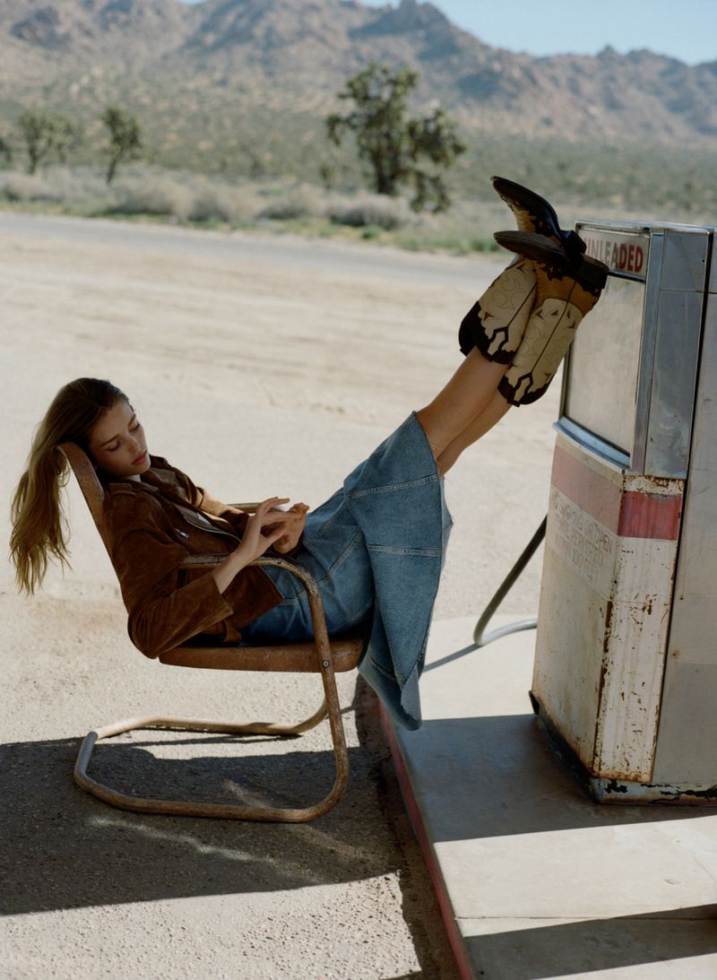 A woman in a brown jacket and jeans reclines on a metal chair next to an old gas pump in a sunlit desert landscape with mountains.