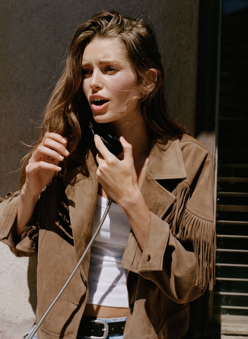 Young woman in fringed suede jacket using vintage telephone outdoors against wall, warm tones, casual style, natural light.