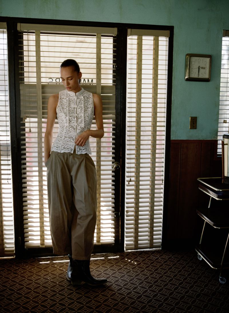 A slender woman with a delicate white floral top and khaki trousers stands in front of a window with venetian blinds inside a room.