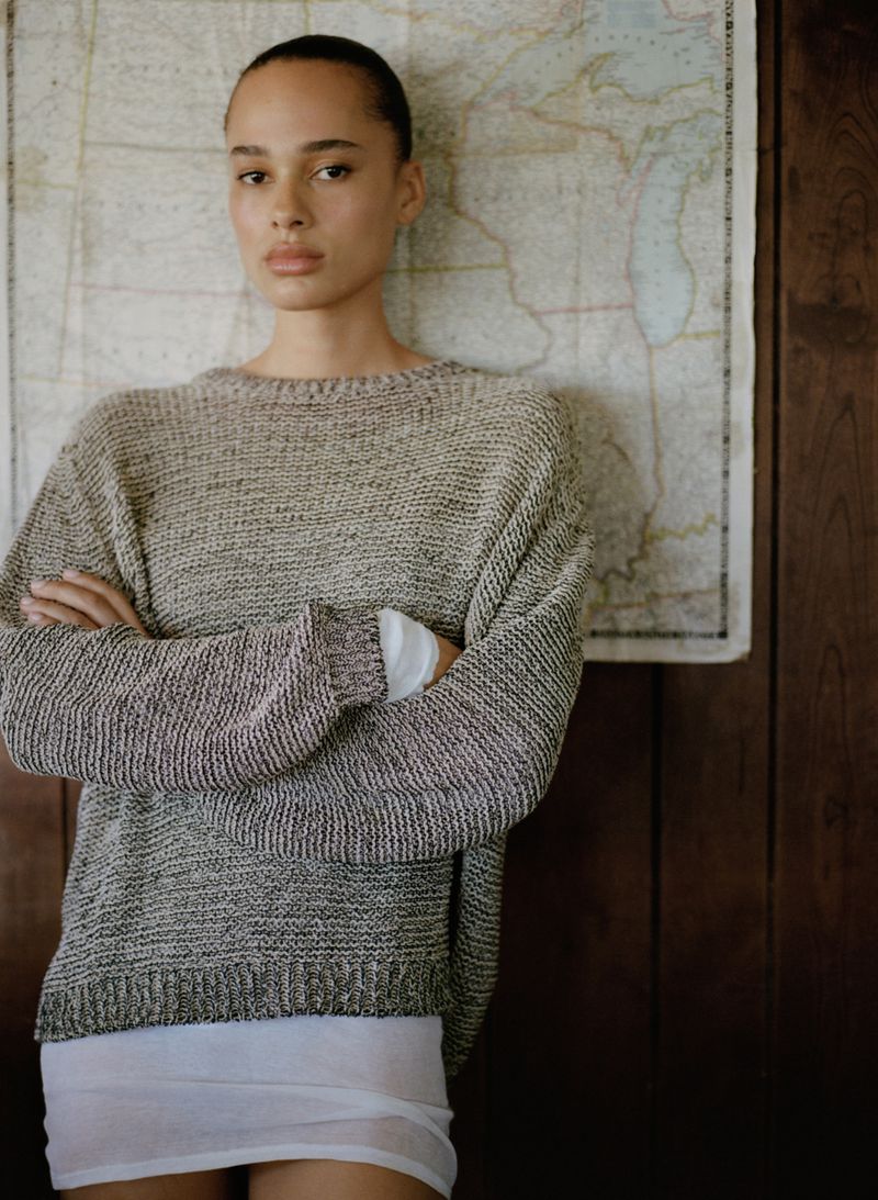 A woman with her hair pulled back wears a beige knit sweater and white undershirt, posing before a vintage map on a wood-paneled wall.