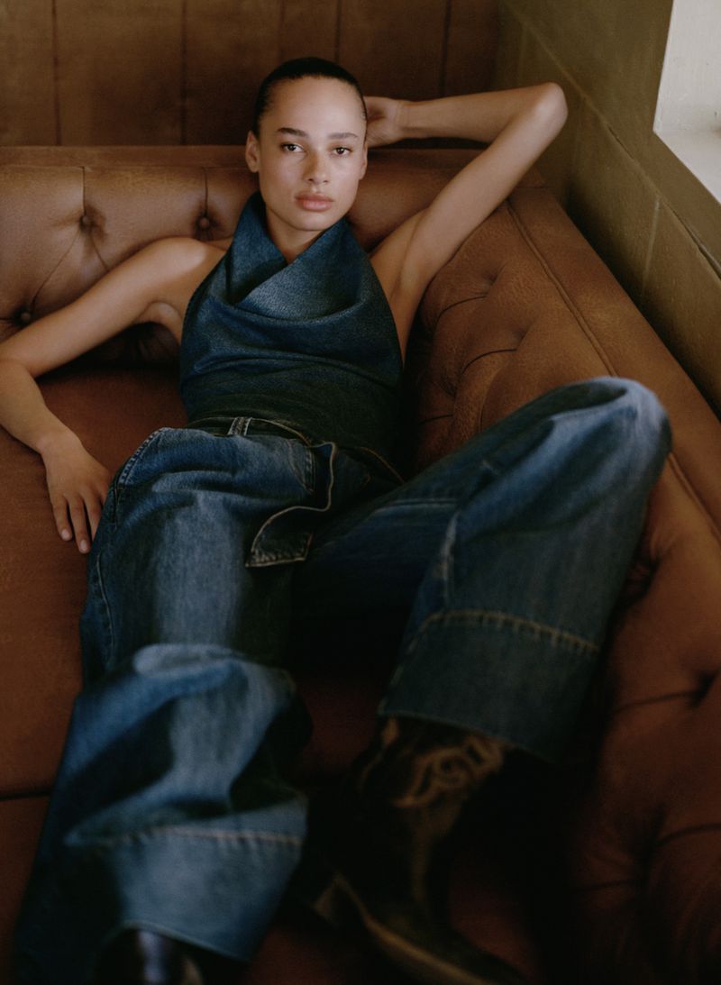 A model is reclining on a brown leather couch, wearing a blue denim halter top and blue jeans, with black boots.