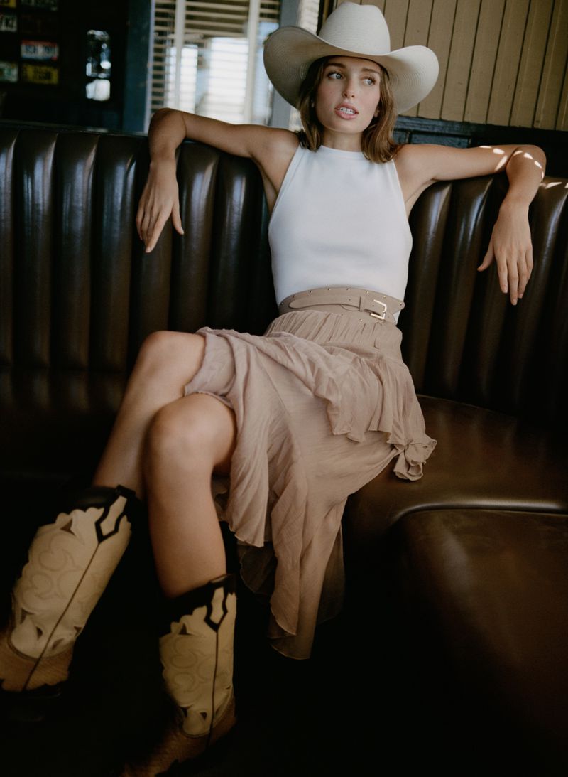 A young woman with long brown hair wearing a white tank top, beige skirt, cowboy hat, and cowboy boots sits on a brown leather couch.