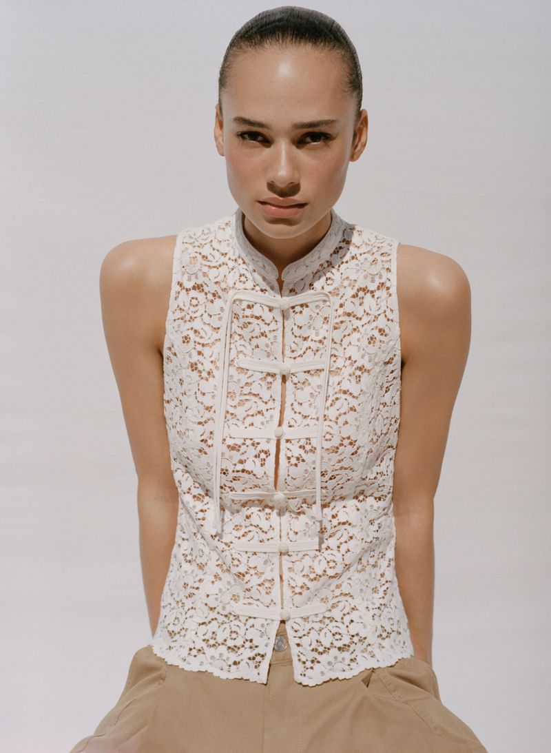Portrait of an adult woman wearing a white lace top with a high collar, complemented by khaki pants, set against a clean white background.