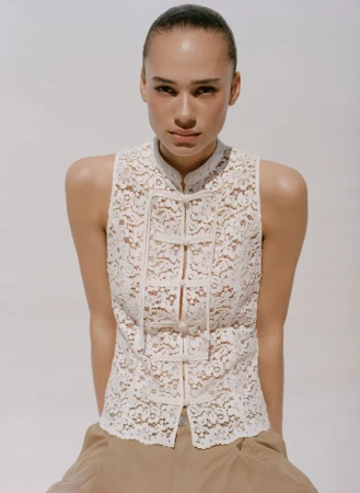 Portrait of an adult woman wearing a white lace top with a high collar, complemented by khaki pants, set against a clean white background.