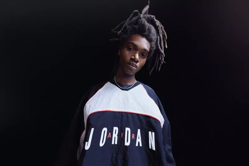Portrait of a young Black man with dreadlocks wearing a Jordan brand sweatshirt against a dark background in a studio setting.