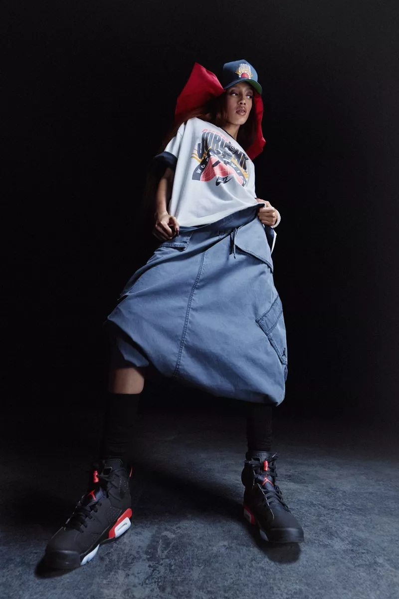 Woman in streetwear posing in a studio wearing an oversized skirt, graphic t-shirt, ball cap, and black and red high-top sneakers.