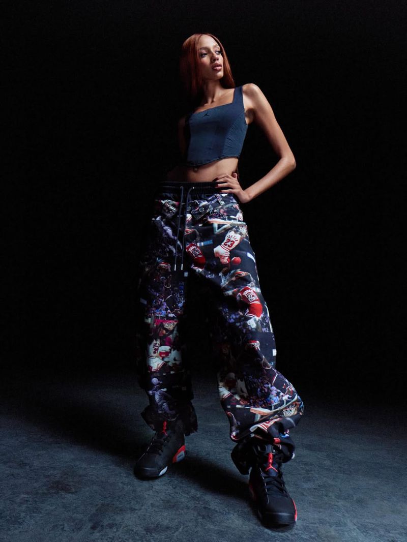 A young adult woman stands confidently in a denim top and printed pants, posing against a dark background in a fashion editorial style.