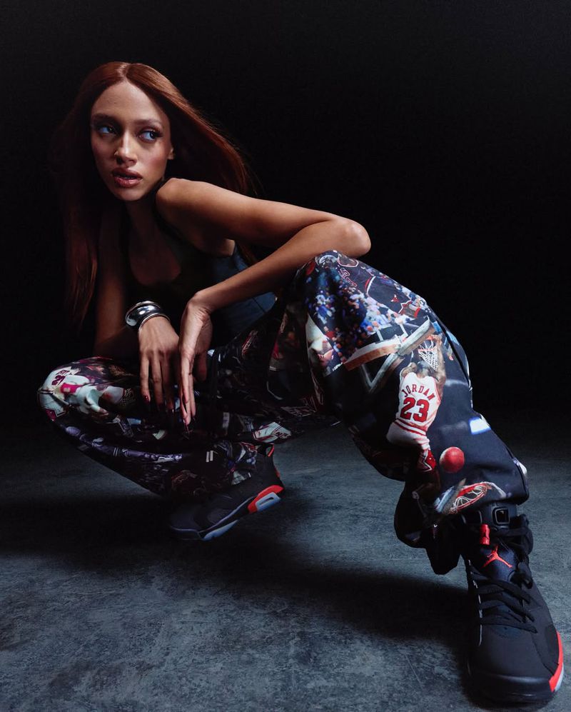 Female model crouches in patterned pants and black sneakers in a studio. The photo has a dark background and medium shot.