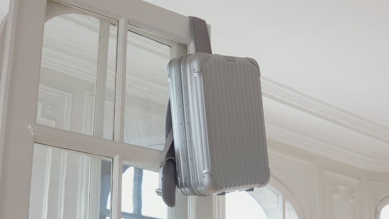 Silver carry-on suitcase hanging from a white door with glass panels, minimalist design, indoor setting, neutral color palette, and soft lighting.