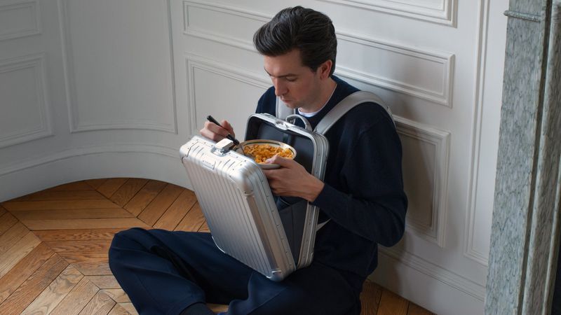 Man in navy sweater and grey backpack sitting on herringbone floor with metal suitcase open, eating out of bowl near wall.