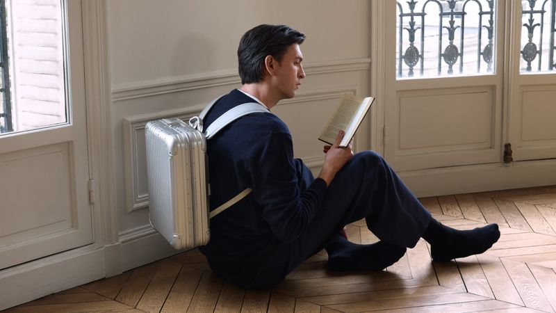 Man seated indoors reading a book with a silver suitcase attached to his back with straps, light streaming through a window.