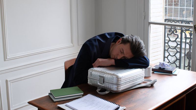 Tired man in suit rests head on silver suitcase at wooden desk with books and papers. Interior shot near window with natural light.