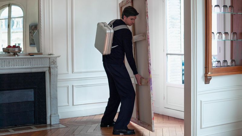 Adult male carries painting in room with backpack. Person transports large painting through doorway, white walls and herringbone floor.
