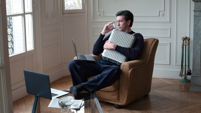 Adult male sits in an armchair in a bright living room with laptops, talking on a smartphone, and holding a silver suitcase.