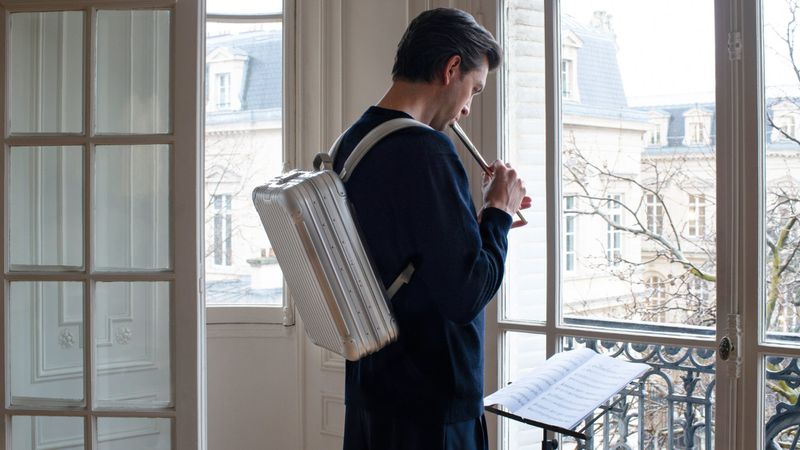 Adult male plays the flute in front of a window with a building view, wearing a backpack and standing near sheet music.