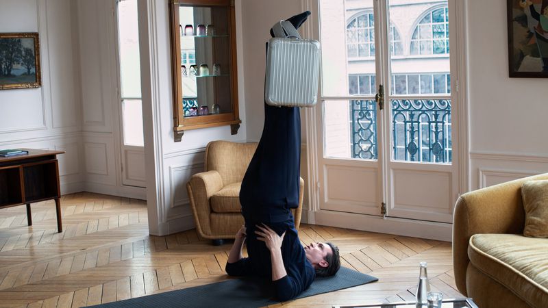 Adult is doing yoga in a home interior with a suitcase on her legs, hardwood floors, and large windows with natural light.