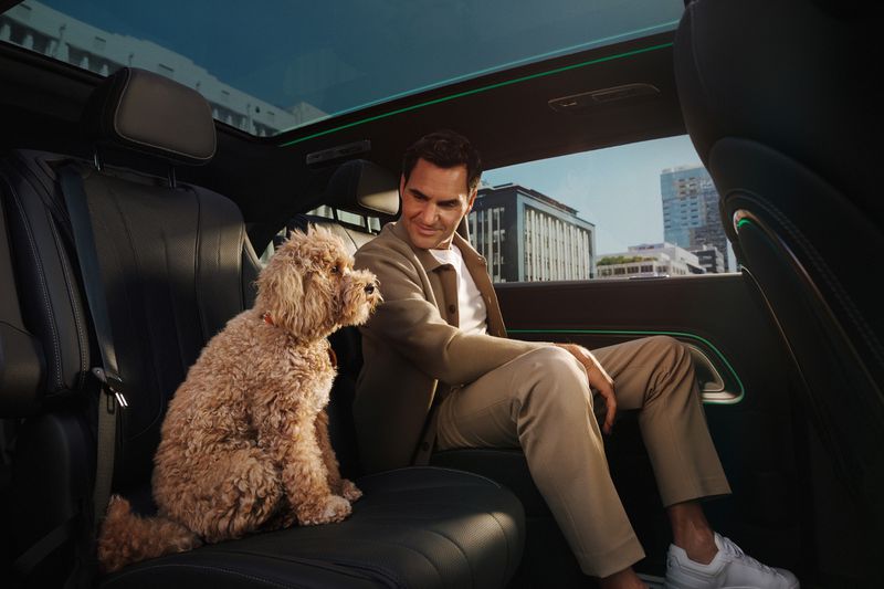 Roger Federer with a Goldendoodle dog sitting in a luxury car, leather interior, and city buildings visible through the sunroof.