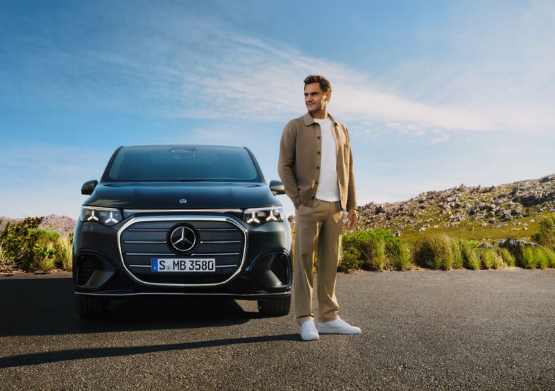 Roger Federer poses with a dark grey Mercedes EQV van against a bright blue sky and green landscape on a sunny day.