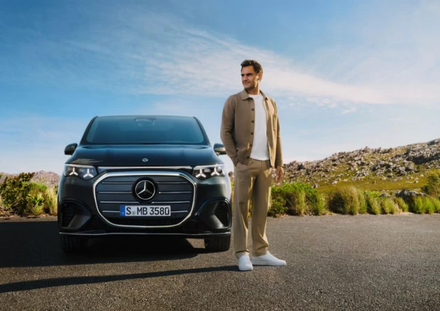 Roger Federer poses with a dark grey Mercedes EQV van against a bright blue sky and green landscape on a sunny day.