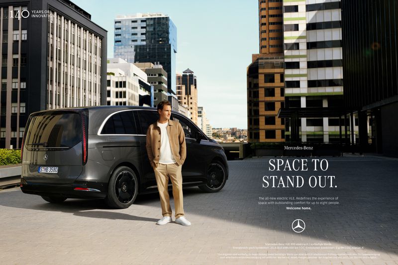 Man poses with a dark gray Mercedes-Benz VLE in an urban setting. Buildings rise in the background under a clear, blue sky.