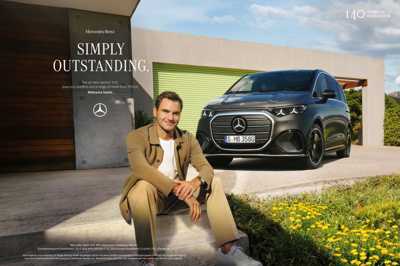 Roger Federer sits on steps near a dark grey Mercedes-Benz electric VLE car outside a modern house with green and yellow flowering ground cover.