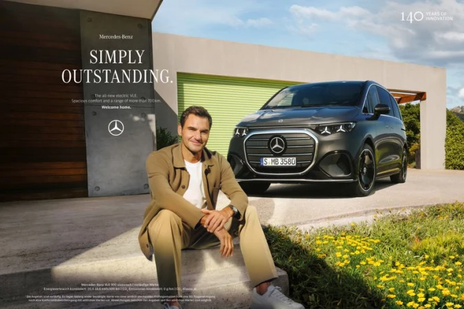Roger Federer sits on steps near a dark grey Mercedes-Benz electric VLE car outside a modern house with green and yellow flowering ground cover.