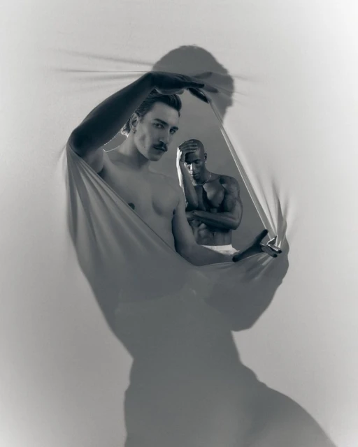 Black and white portrait of two adult men partially visible within a stretched piece of fabric, indoor studio setting, artistic.