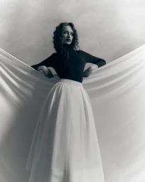 Monochrome fashion portrait of a woman with dark curly hair wearing a long skirt, posed against draped fabric backdrop
