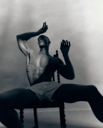 Shirtless Black man seated on a carved wooden chair with arms raised against an overcast sky