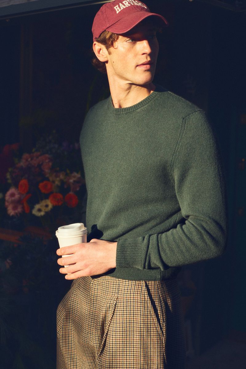 A young adult male in a sweater and cap holds a coffee cup in an urban setting with a background of flowers.