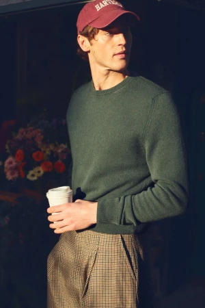 A young adult male in a sweater and cap holds a coffee cup in an urban setting with a background of flowers.
