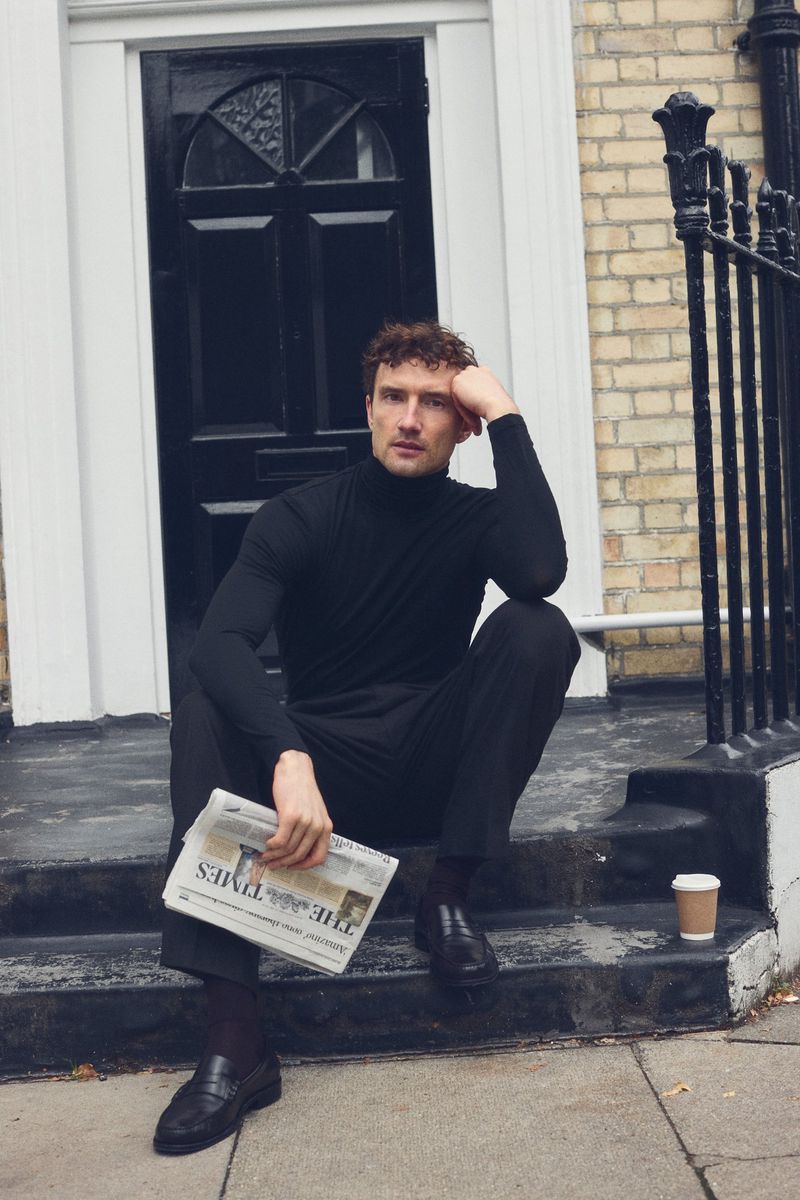 Man in black turtleneck sitting on stairs with newspaper and coffee cup outside a building with a black door and brick wall.