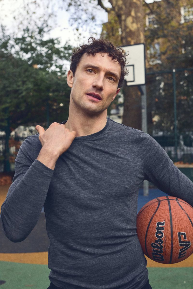 Adult male poses with a basketball on an outdoor court with trees in the background, wearing a grey long-sleeved shirt. Basketball hoop visible.