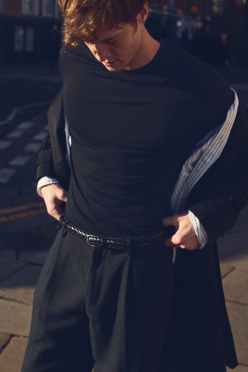 Adult man in a black shirt and wide pants adjusts his belt on a sidewalk against a backdrop of buildings in an urban environment.