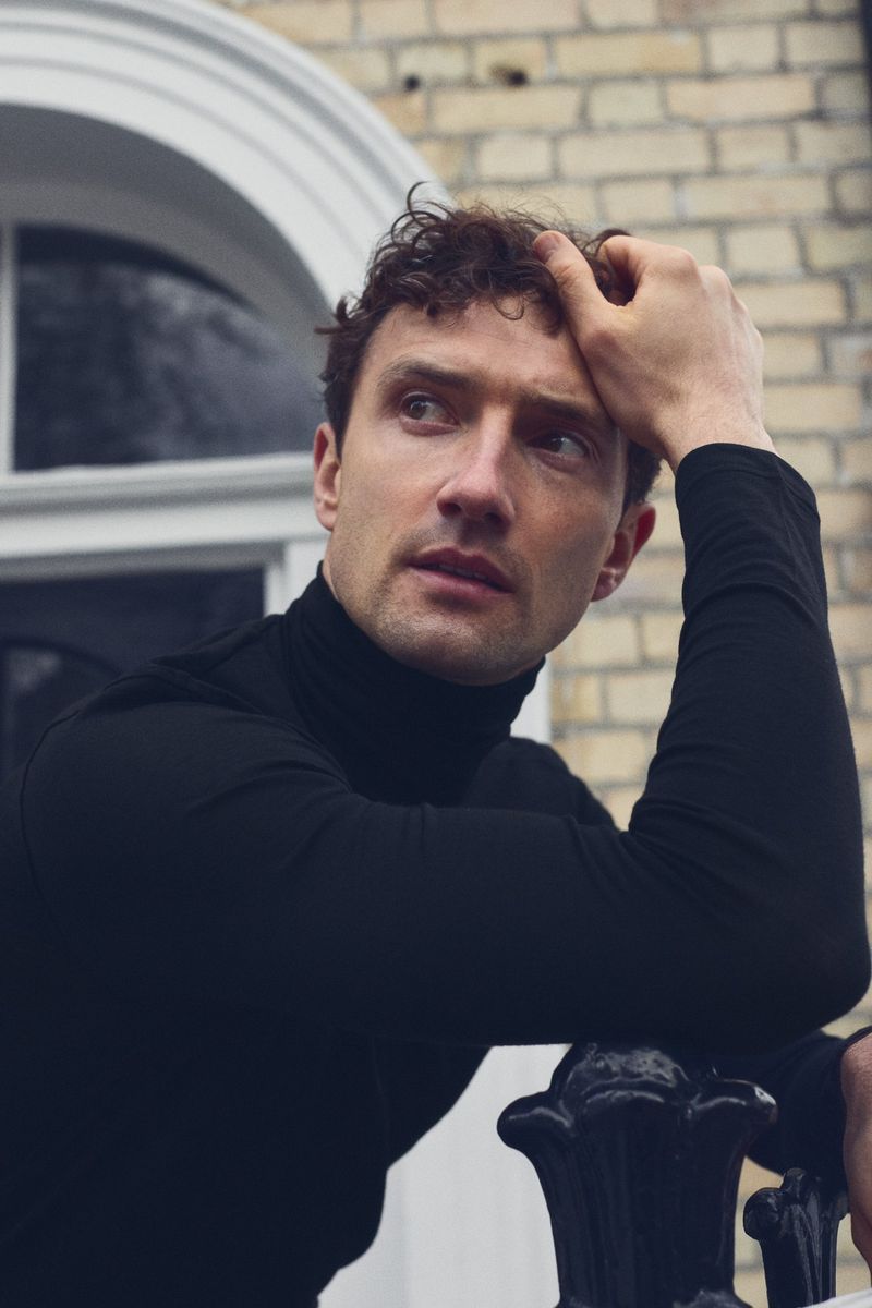 Portrait of a handsome adult man with brown curly hair in a black turtleneck leaning on a black wrought iron fence outdoors.